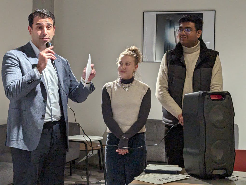 A person speaks into a microphone, holding paper, flanked by two attentive people in a room with a speaker and a framed picture on the wall.