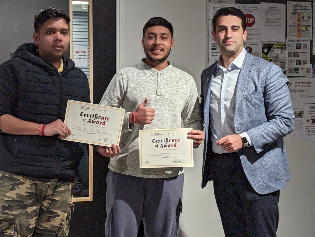 Three people stand indoors, two holding certificates of award. The middle person gives a thumbs-up, and they all appear proud.