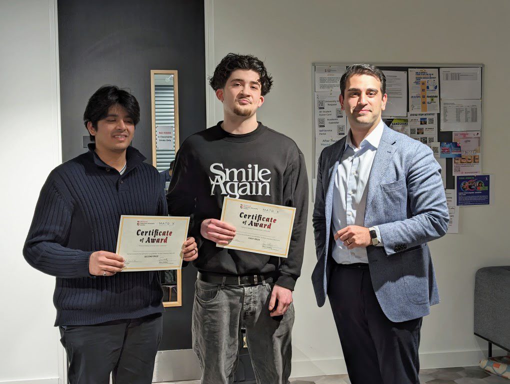 Three people are standing indoors, smiling, and holding certificates of award. One is wearing a blazer, another a slogan sweatshirt, and the third a sweater.