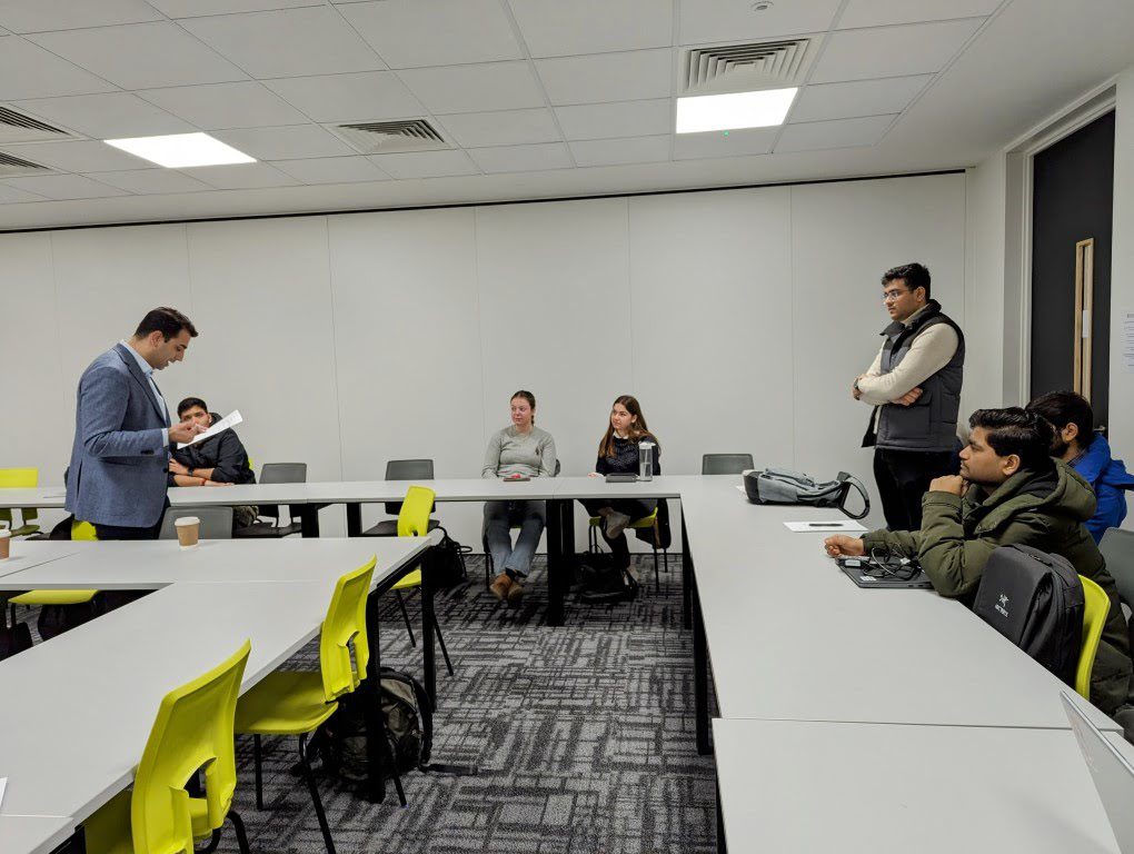 The image shows a modern classroom setting with one standing person appearing to address seated individuals, some with attentive expressions, in a casual atmosphere.